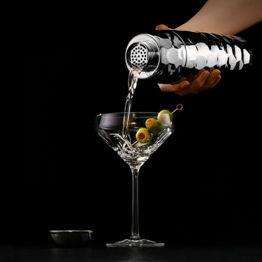 Bartender pouring a cocktail into a martini glass with olives on a black background