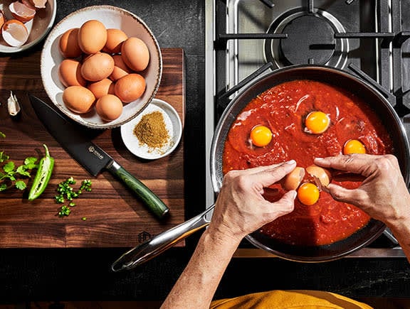 Person cracking eggs into a pan of simmering tomato sauce on a stove, surrounded by ingredients.