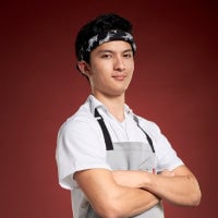 A confident chef in a white uniform and apron, standing against a red background.