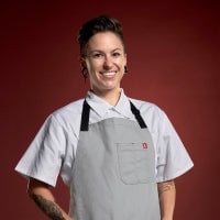 A smiling individual in a chef's uniform with a red background.